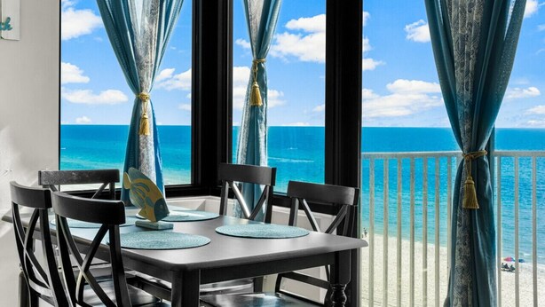 A balcony with a view of the ocean, a table with chairs, and blue curtains tied with tassels.