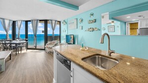 A kitchen with a granite countertop, stainless steel sink, and a view of the ocean through large windows.