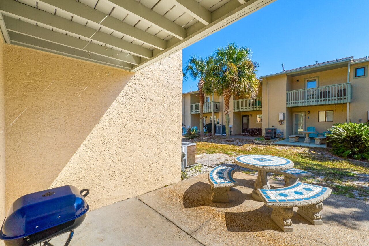 A covered patio with a barbecue grill, outdoor seating, and a view of a residential building with balconies and palm trees.