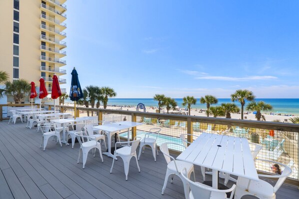 Una terraza con mesas y sillas blancas, con vista a una playa con palmeras y una piscina.