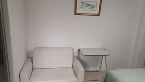 A small, clean room with a white sofa, a small table, and a bed with a green blanket. - Shin Awaji Building II 202  (Osaka)