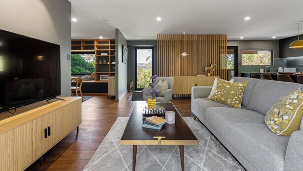 Ein modernes Wohnzimmer mit einem Flachbildfernseher, einer Holz-Unterhaltungsanlage, einem grauen Sofa mit dekorativen Kissen, einem Couchtisch mit Büchern und einer Vase sowie einem Blick ins Freie durch ein großes Fenster.