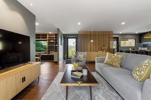 A modern living room with a flat-screen TV, a wooden entertainment unit, a grey sofa with decorative pillows, a coffee table with books and a vase, and a view of the outdoors through a large window. - Equinox - Modern Highlands Charm in Mittagong (Mittagong)