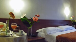 Une chambre d’hôtel avec un lit parfaitement fait, une table de chevet sur laquelle se trouvent un verre de jus d’orange, une tasse et une assiette avec un dessert. Il y a deux vases avec des fleurs sur la table.