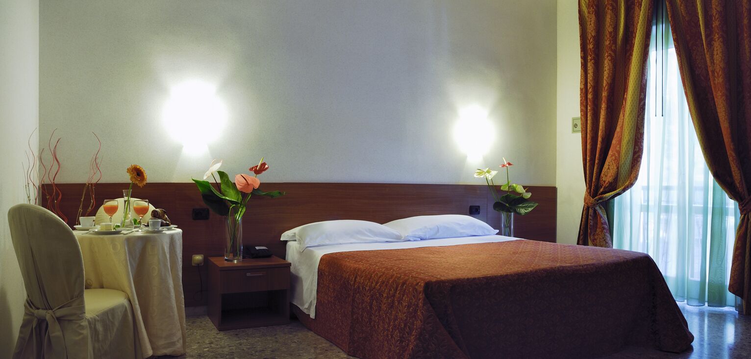 Une chambre d’hôtel avec un lit, une table de chevet, une chaise, une table ornée d’un vase de fleurs et une fenêtre avec des rideaux.