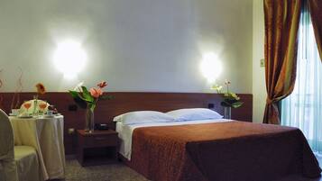 Une chambre d’hôtel avec un lit, une table de chevet, une chaise, une table ornée d’un vase de fleurs et une fenêtre avec des rideaux.
