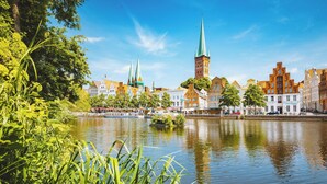 Udsigt over en flod med historiske bygninger, et markant tårn og spir, omgivet af frodig vegetation.