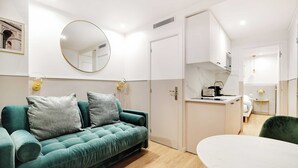 Una habitación moderna con un sofá azul verdoso, un espejo redondo, una mesa de comedor blanca y una kitchenette.