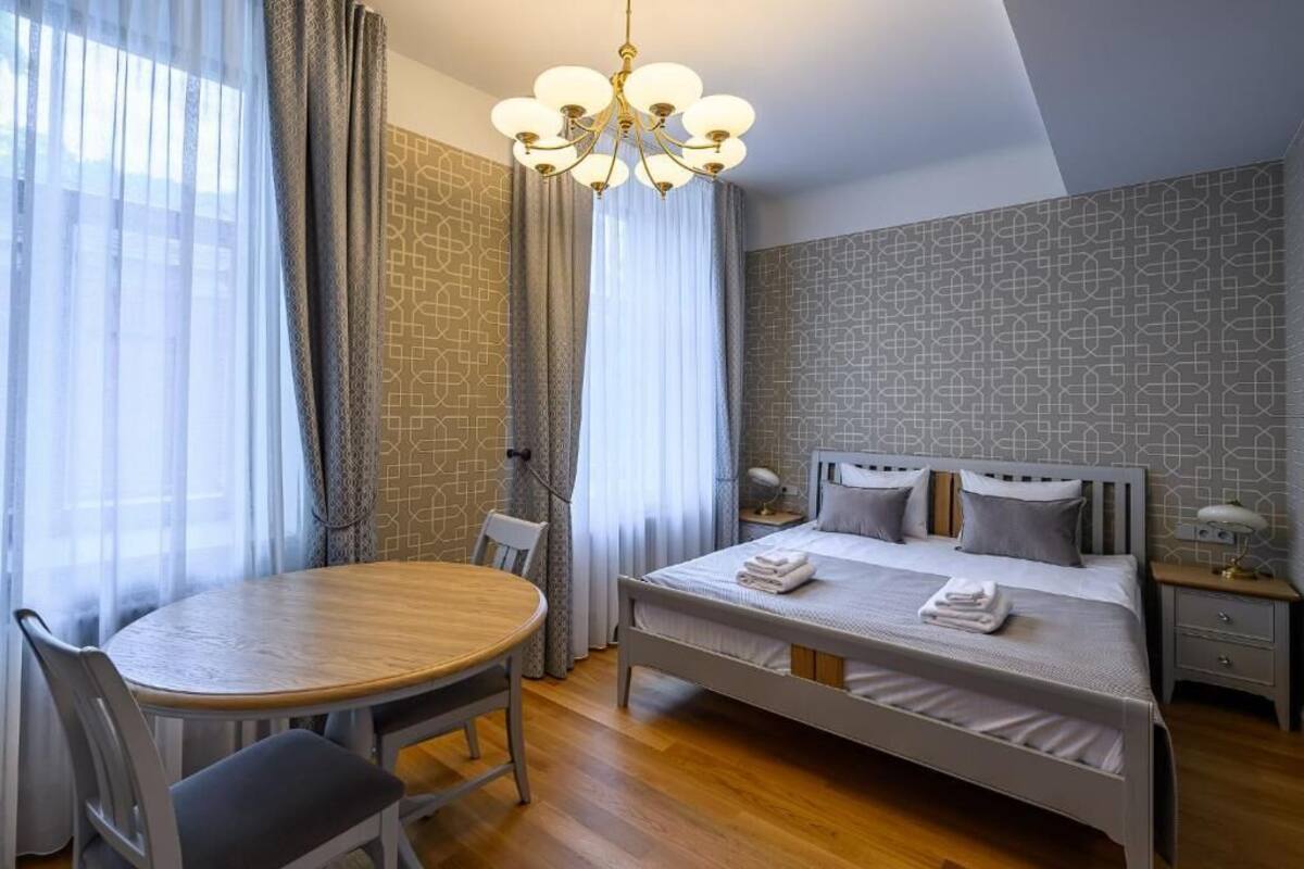 Una habitación de hotel con cama, una mesa de comedor redonda, sillas, una araña y cortinas.