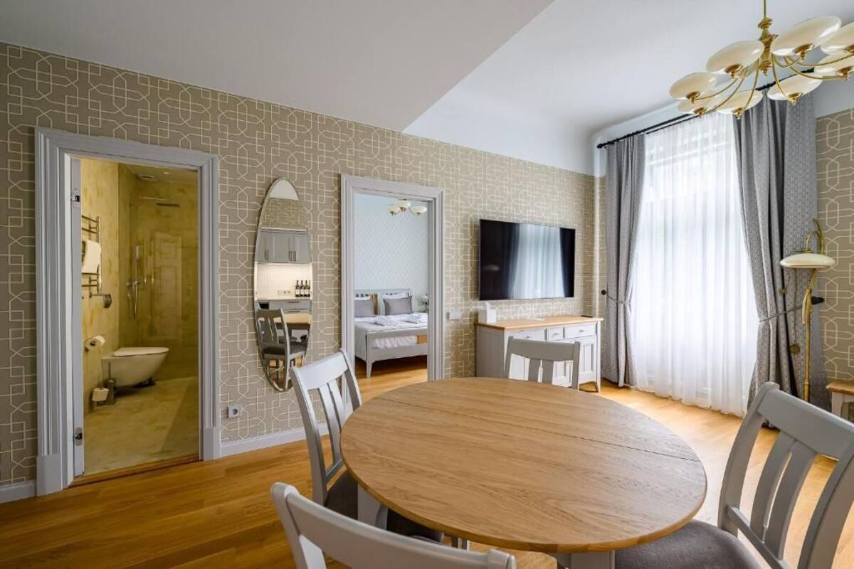Una habitación de hotel moderna con una mesa de comedor de madera, sillas blancas, un televisor y un baño visible a través de una puerta abierta.