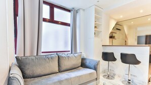 Een moderne woonkamer met een grijze bank, twee zwarte barkrukken, een glazen salontafel en een keuken in de achtergrond.