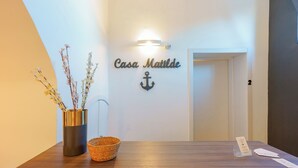 Un comptoir d’accueil avec une enseigne indiquant « Casa Maitde » et un symbole d’ancre. On y trouve un vase avec des fleurs et un panier.