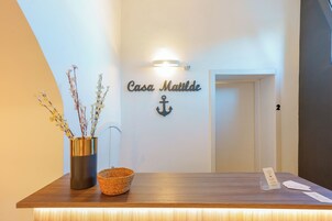 Una recepción con un letrero que dice "Casa Maitde" y un símbolo de ancla. Hay un jarrón con flores y una cesta en el mostrador.