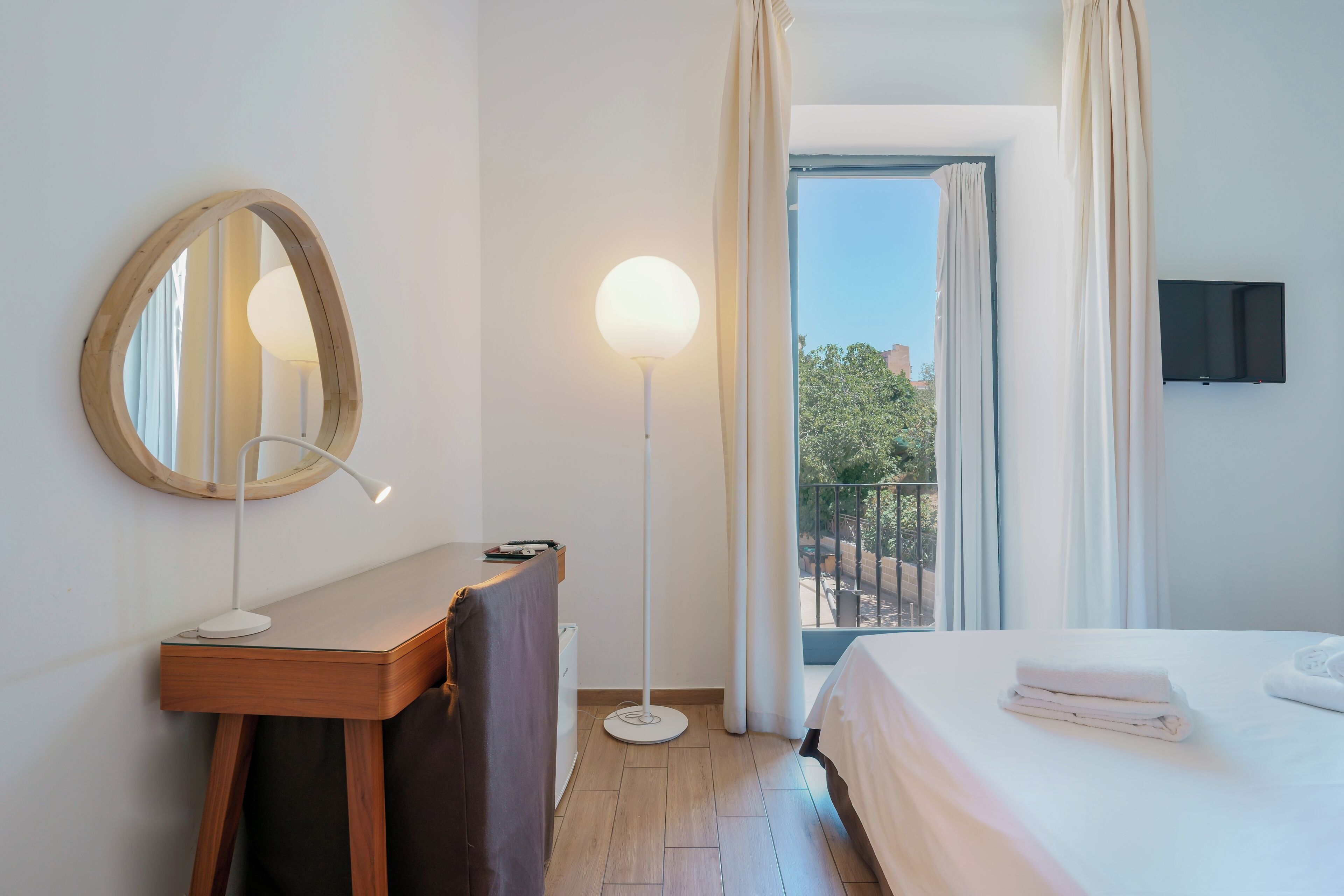 A hotel room with a bed, a desk with a lamp, a mirror, a flat-screen TV, and a view of the outdoors.