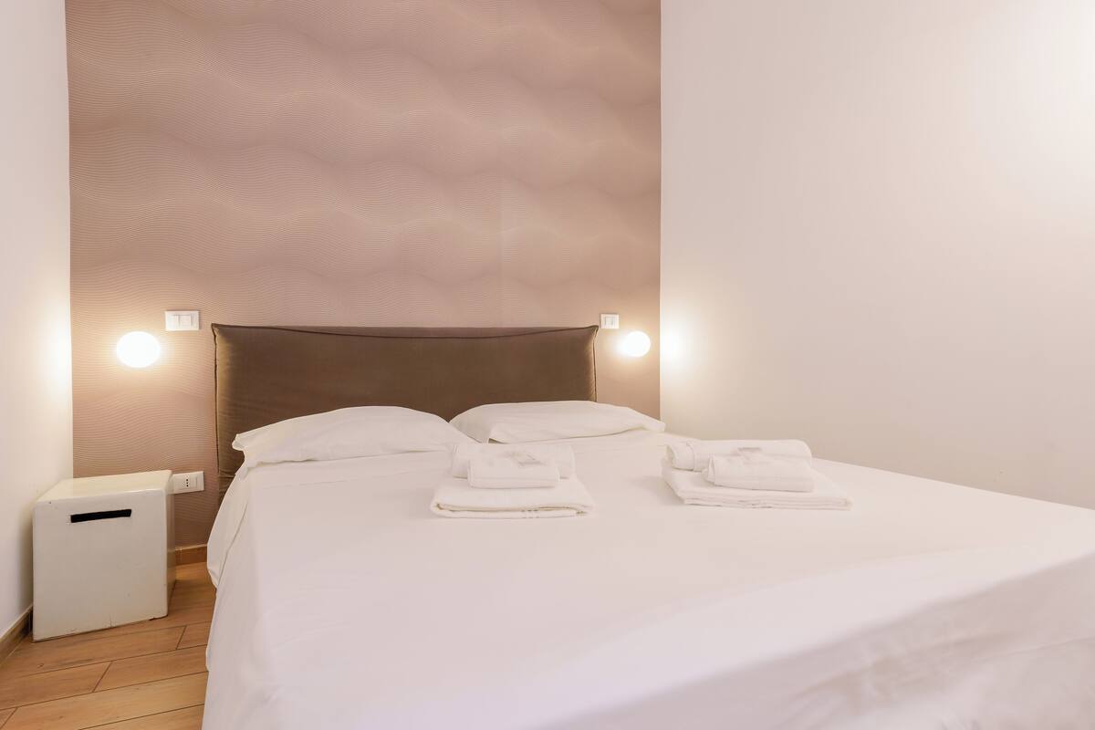 A neatly made bed with white linens and towels, a bedside table, and wall-mounted lights.