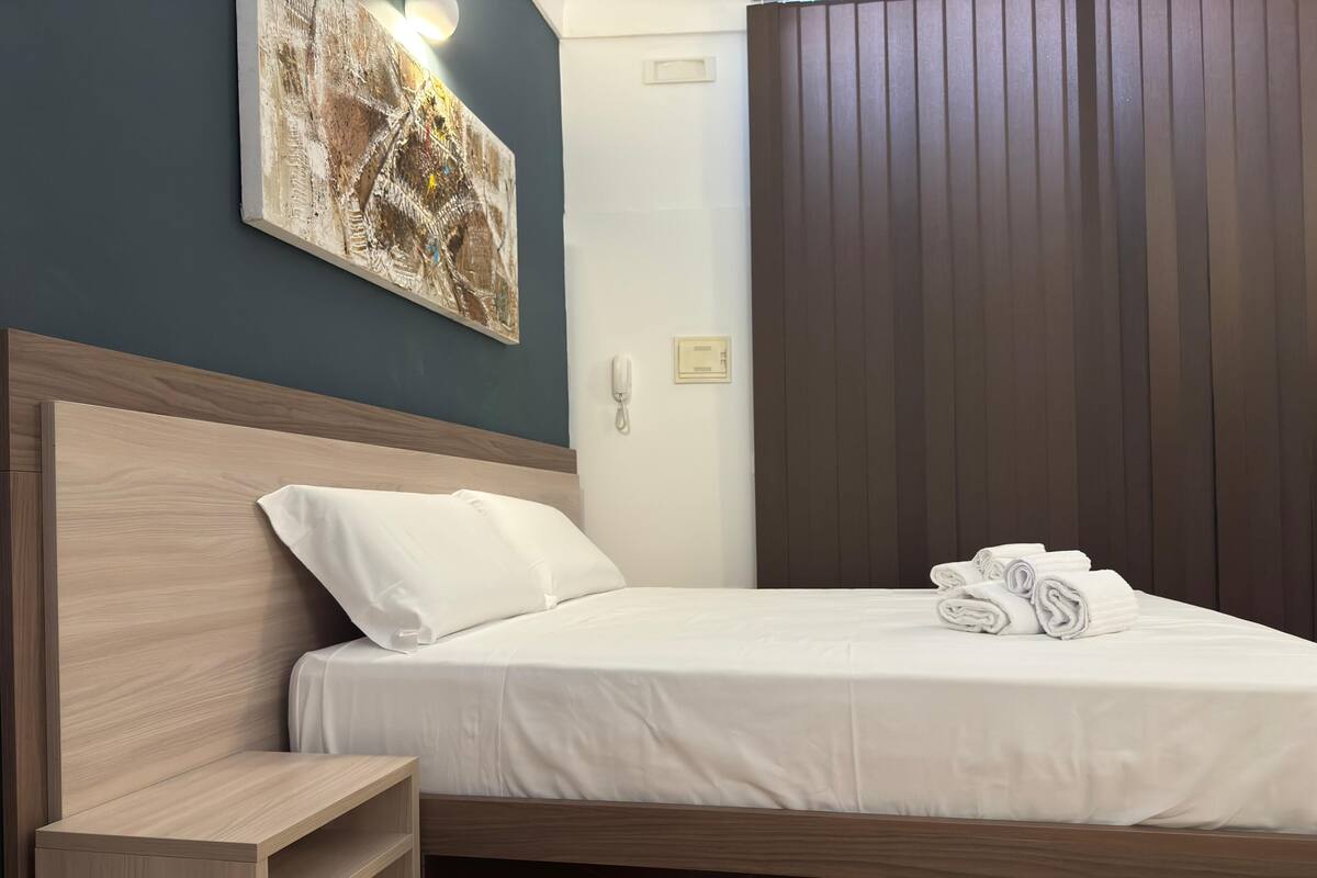 A neatly made bed with white linens and rolled towels, a wooden headboard, a bedside table, and a wall-mounted phone.