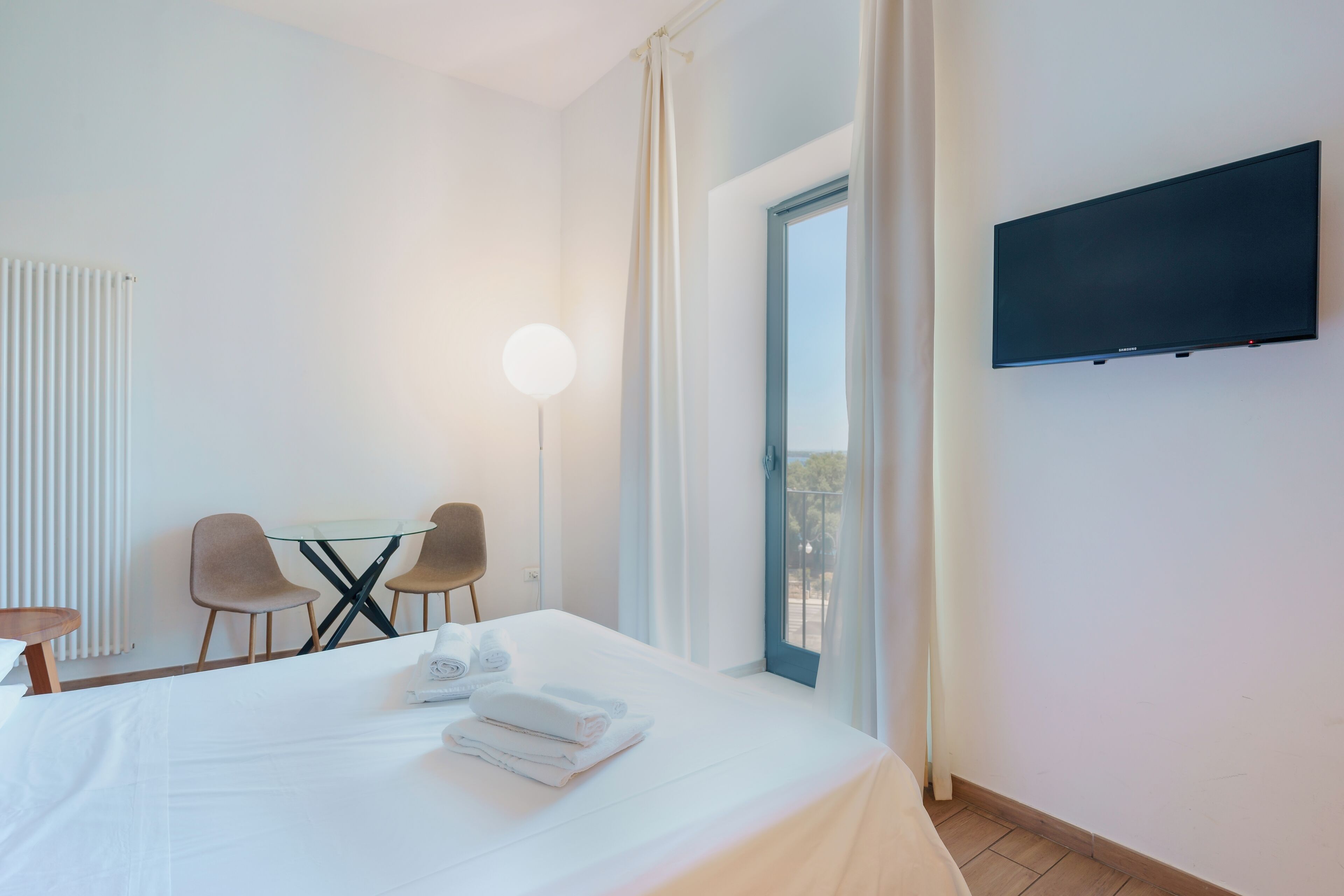 A hotel room with a bed, a desk with chairs, a flat-screen TV mounted on the wall, and a window with curtains.