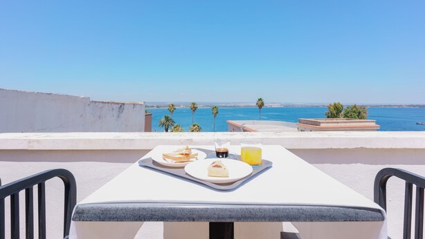 Une terrasse avec une table préparée pour le petit-déjeuner, offrant une vue sur la côte, avec des palmiers et la mer.