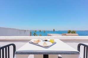A terrace with a table set for breakfast, overlooking a coastal view with palm trees and the sea.