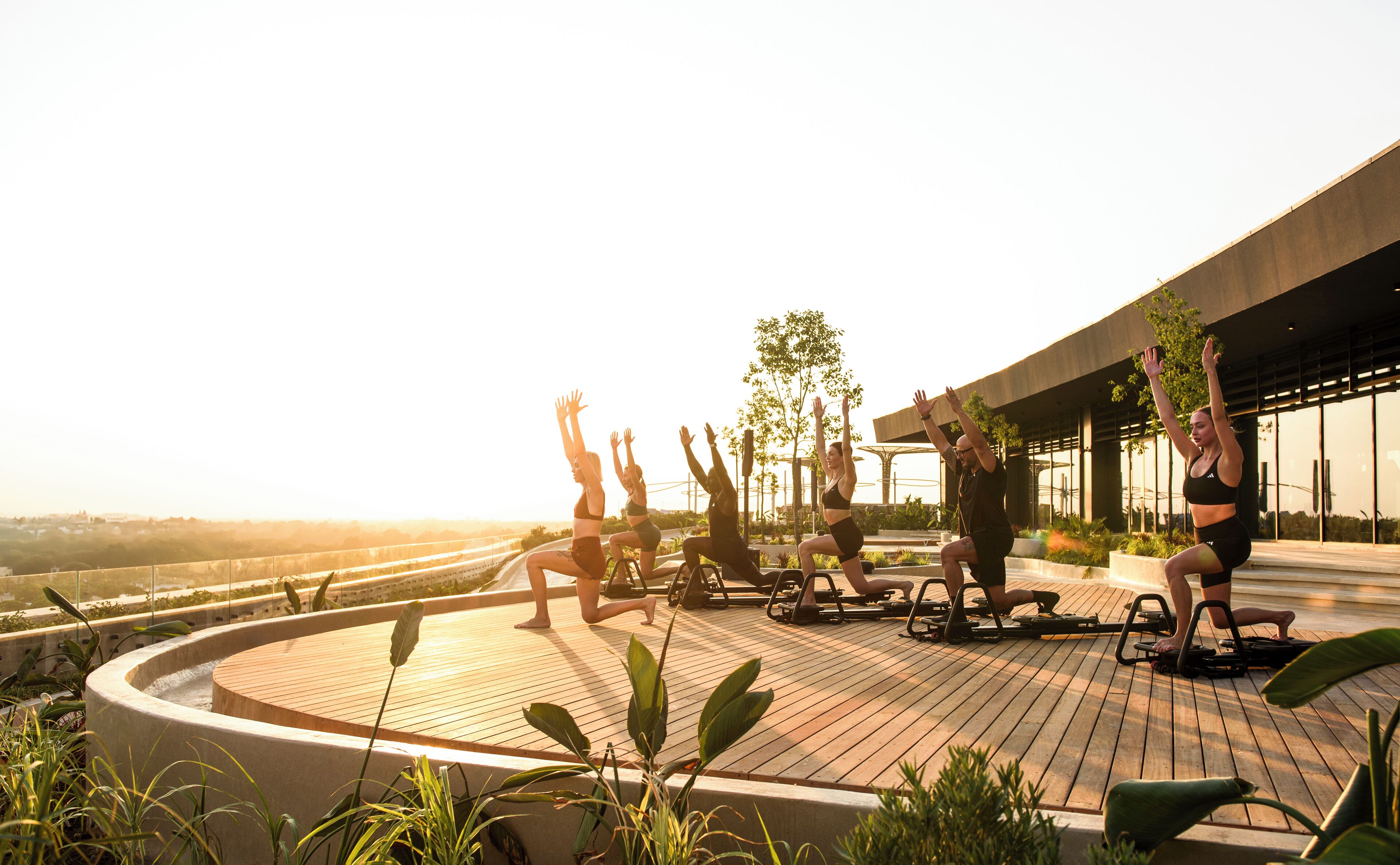 Un groupe de personnes fait de l’exercice en plein air sur une terrasse en bois, équipée de matériel d’entraînement, entourée de verdure et avec un bâtiment moderne en arrière-plan.