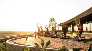 Un groupe de personnes fait de l’exercice en plein air sur une terrasse en bois, équipée de matériel d’entraînement, entourée de verdure et avec un bâtiment moderne en arrière-plan.