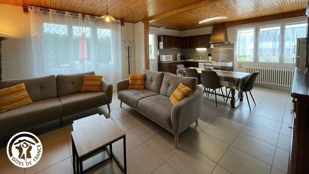 A modern living room with a sofa and armchair, a dining area, and a kitchen.