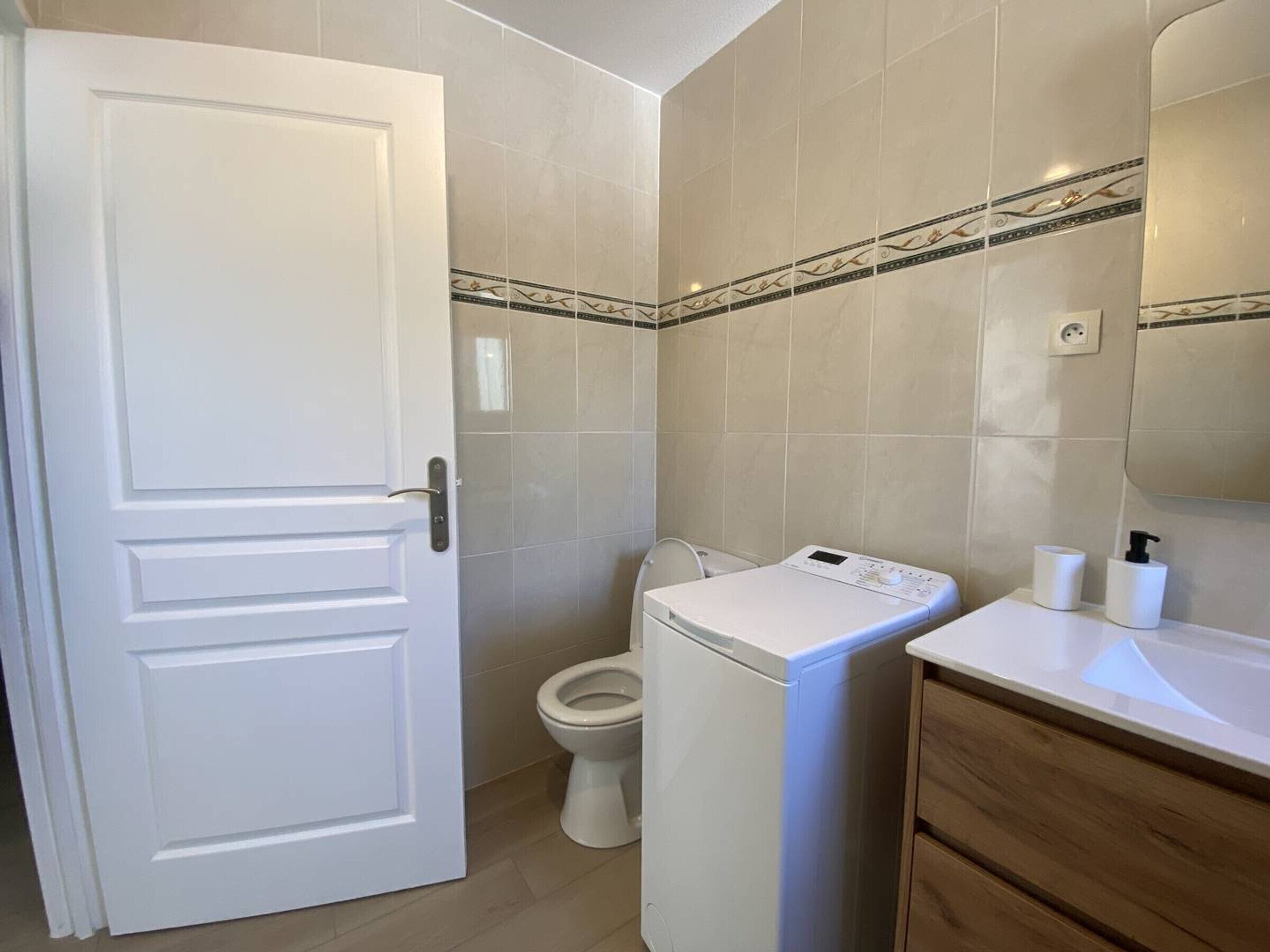 Un baño con puerta blanca, inodoro, lavadora y lavabo con artículos de aseo.