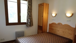 Un dormitorio con cabecera de madera, colcha estampada, ventana con cortinas, armario e iluminación en la pared.