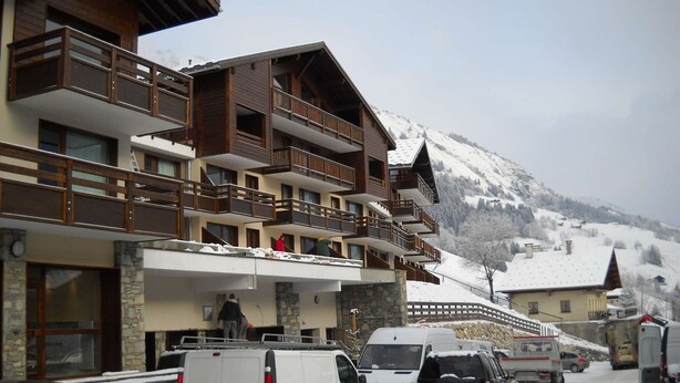 Un edificio de varios pisos con balcones, una montaña nevada al fondo y vehículos estacionados al frente.