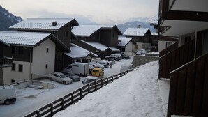 Un pueblo cubierto de nieve con edificios de estilo chalete tradicional, una calle estrecha bordeada de coches aparcados y una vista de montañas lejanas.