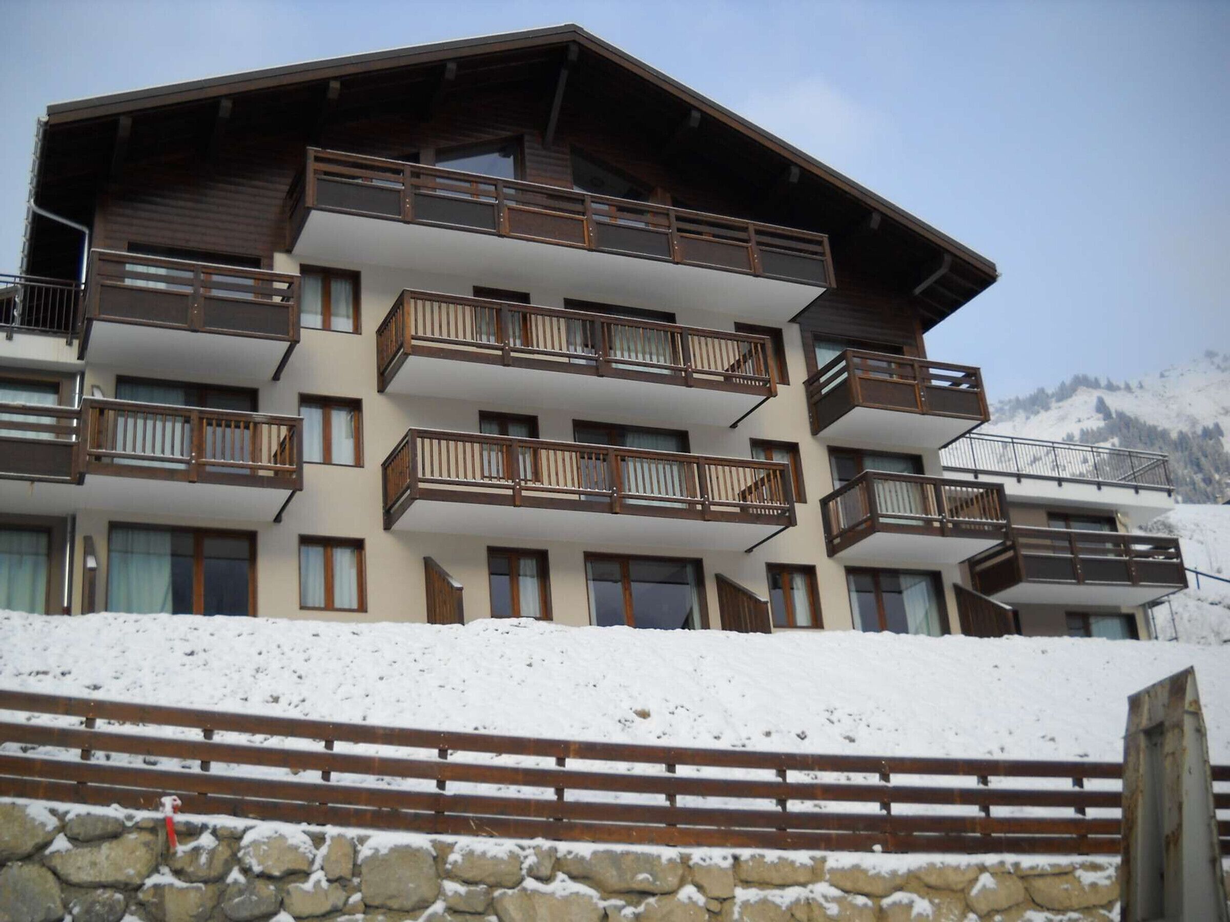 A multi-story hotel with balconies, a stone retaining wall, and a snowy mountain backdrop.