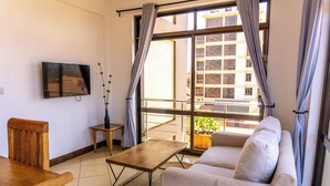 Un salon avec un canapé, une table en bois, un téléviseur fixé au mur et un balcon agrémenté d’une plante.