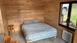Et soveværelse i en træhytte med en seng, en skammel og et vindue med udsigt til en bil udenfor.