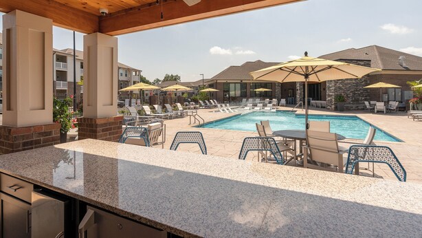 A pool area with lounge chairs and umbrellas, a covered seating area with a ceiling fan, and a view of residential buildings in the background.