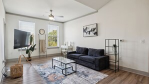 A modern living room with a dark grey sofa, a glass coffee table, and a large flat-screen TV. There are two windows with blinds, a ceiling fan, and a framed abstract painting on the wall.