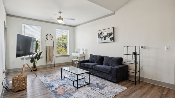 A modern living room with a dark grey sofa, a glass coffee table, and a large flat-screen TV. There are two windows with blinds, a ceiling fan, and a framed abstract painting on the wall. - Overlook 4302 I Stunning 2Bd/2Bth Apt I Pool (Knoxville)
