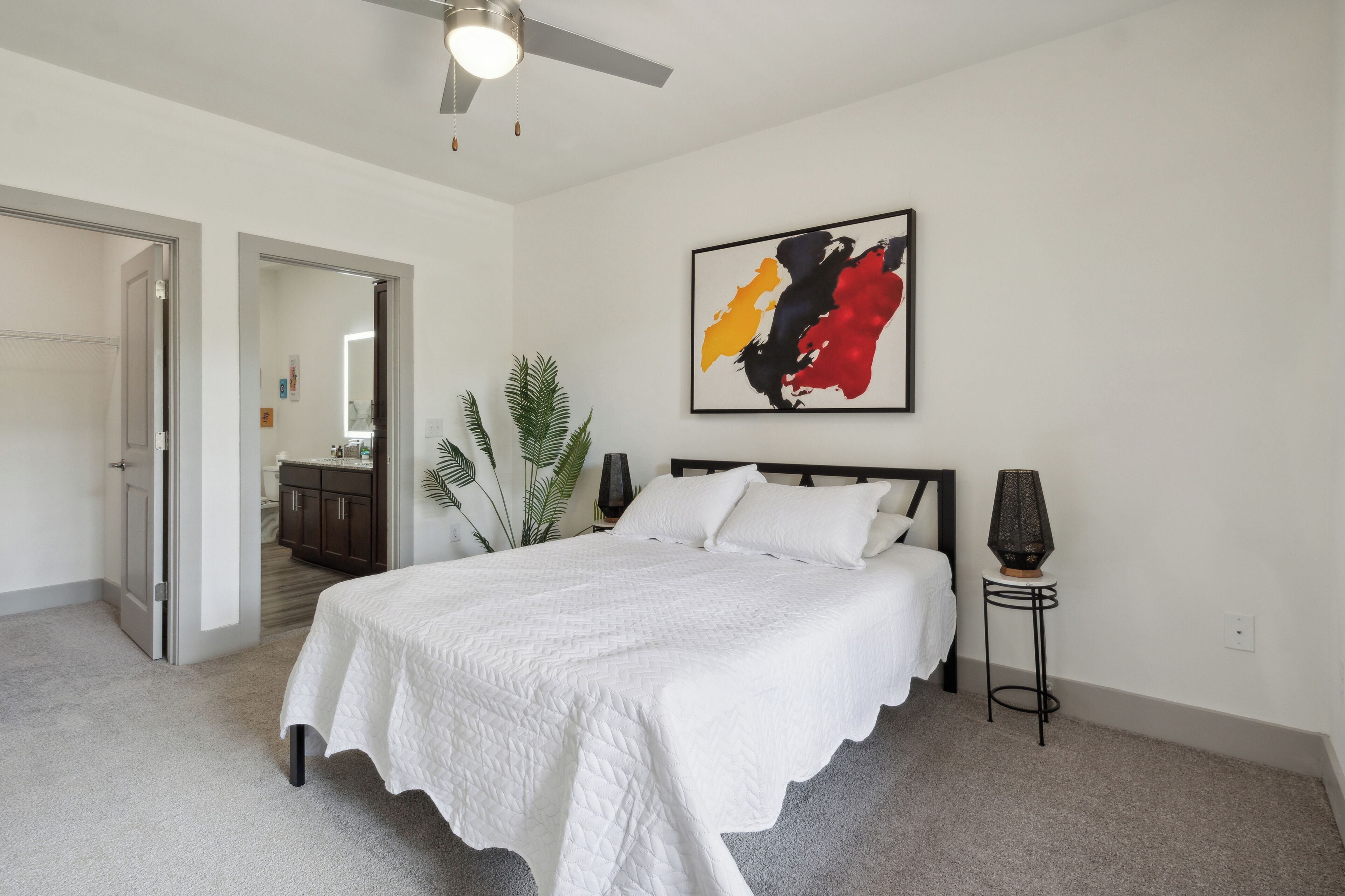 Une chambre à coucher comprenant un lit, un ventilateur de plafond, un tableau abstrait accroché au mur, une table de chevet et une plante en pot.