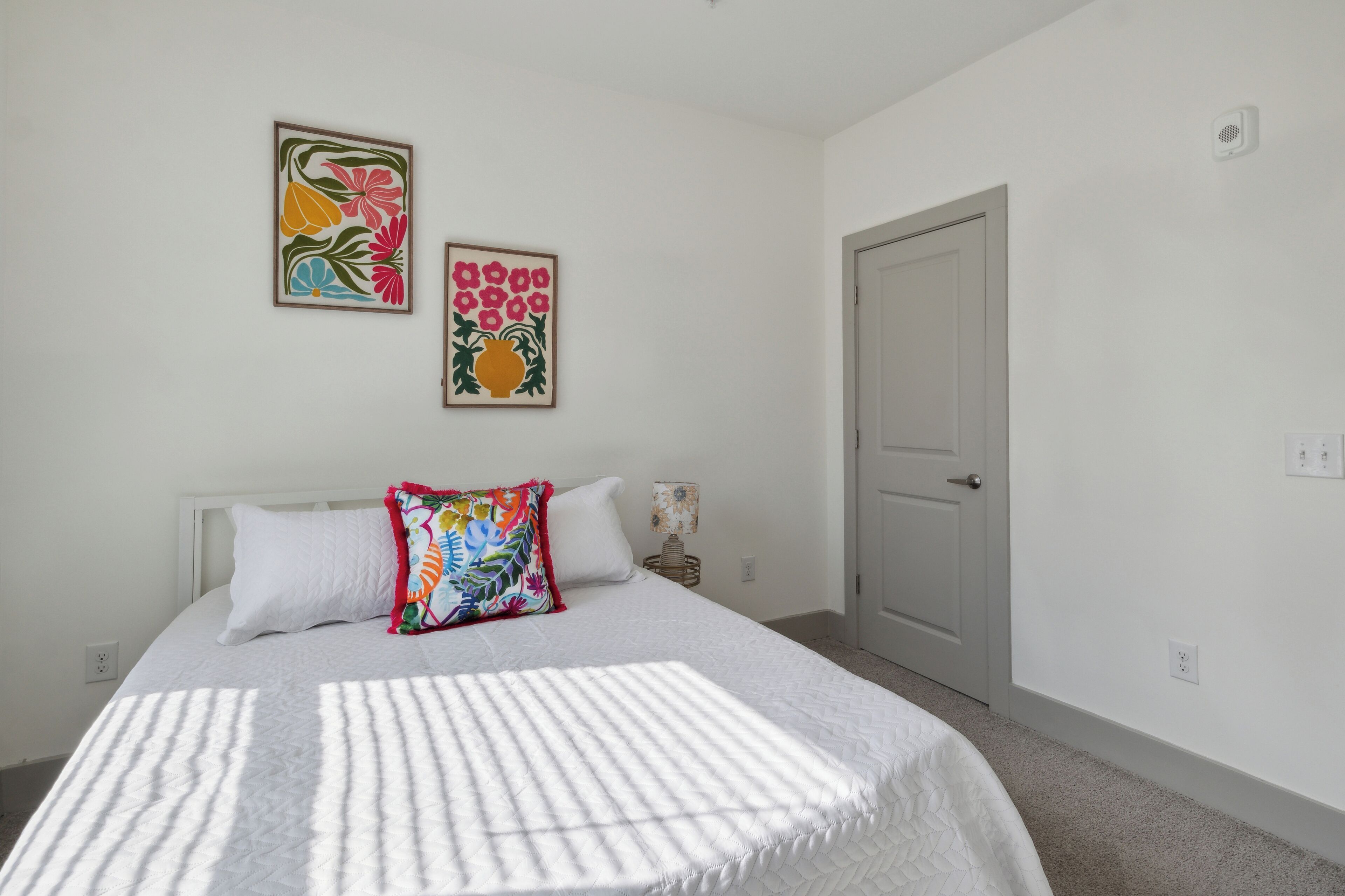 Un dormitorio con cama, paredes blancas, dos cuadros con motivos florales, una puerta y una lámpara de noche.