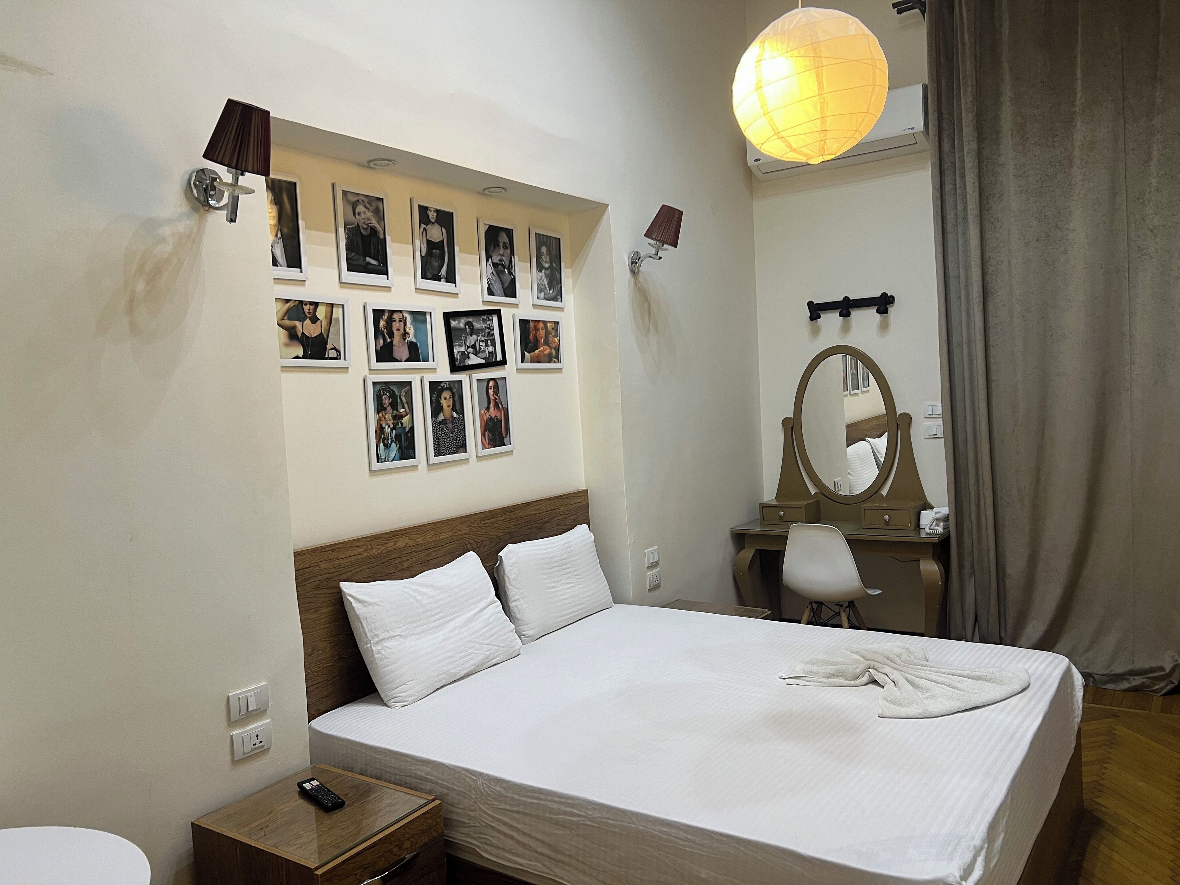 A hotel room with a neatly made bed, a bedside table with a remote control, a wall-mounted mirror, and a series of framed pictures on the wall.