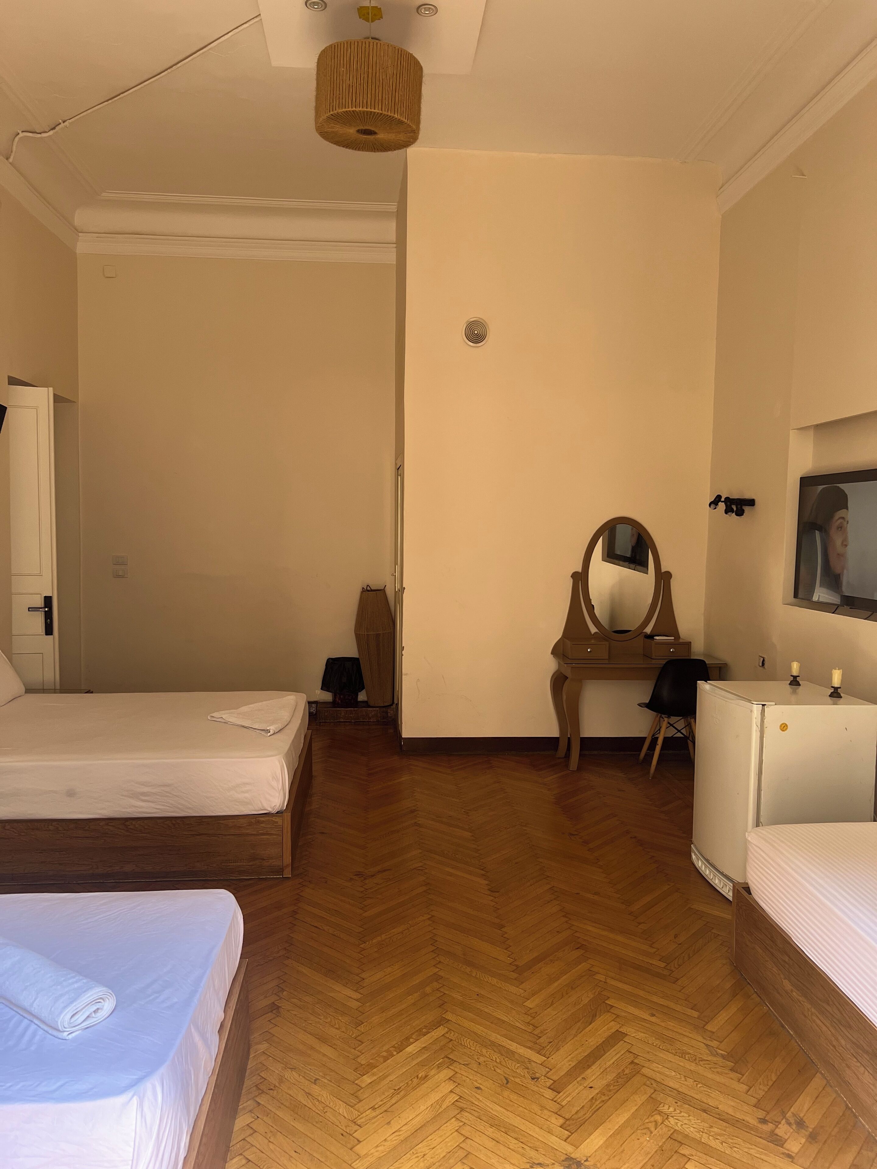 A hotel room with two beds, a wooden herringbone floor, a desk with a mirror, a small refrigerator, and a wall-mounted TV.