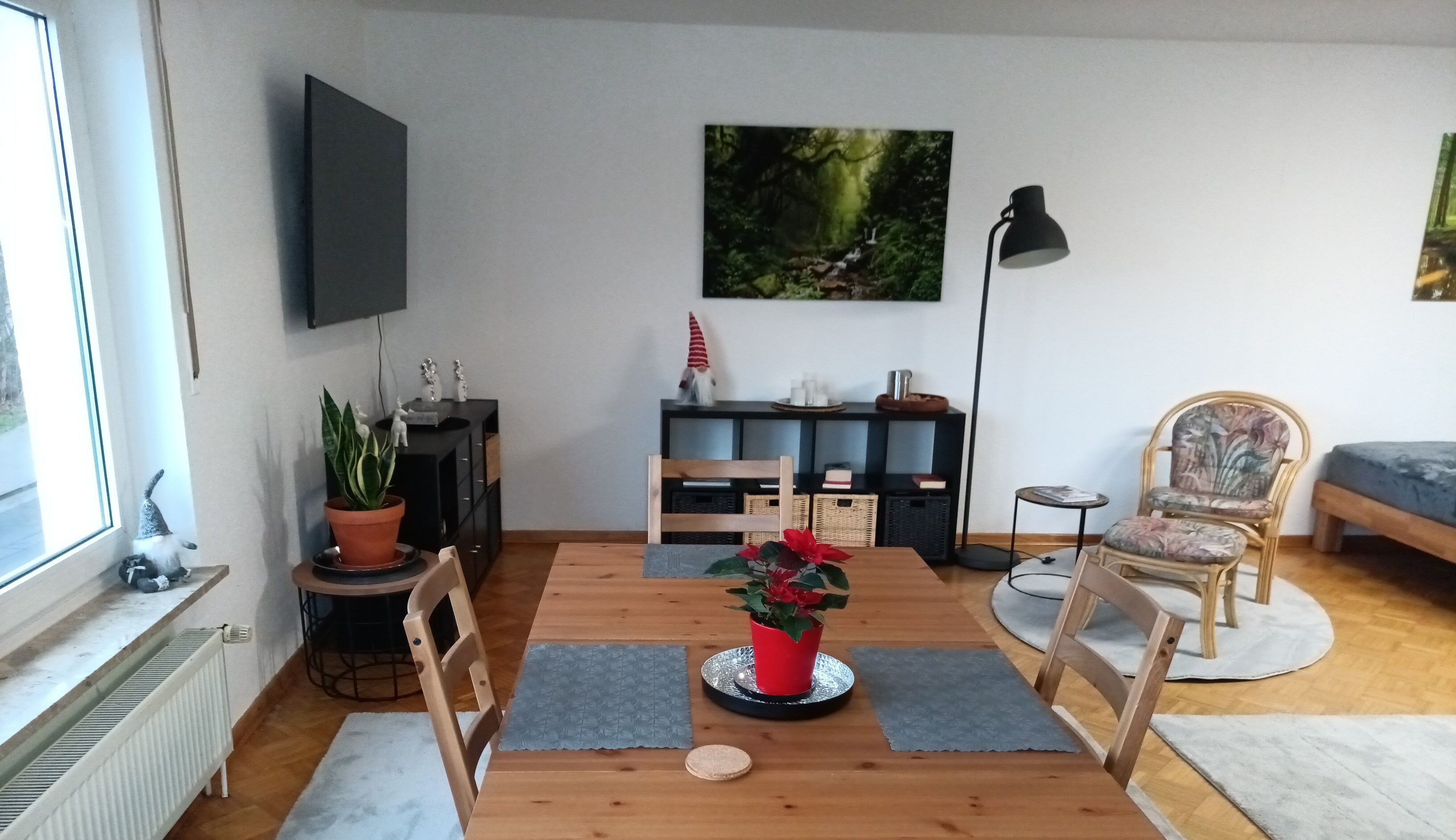 Ein gemütliches Wohnzimmer mit einem Holzesstisch, Stühlen, einem an der Wand befestigten Fernseher, einem Bücherregal und einem großen Fenster.