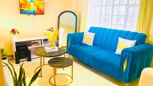 Ein Wohnzimmer mit einem blauen Sofa, einem kleinen runden Couchtisch, einem an der Wand montierten Fernseher und einem Fenster mit Vorhängen.