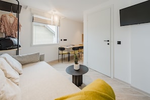 A modern hotel room with a sofa, a dining table with chairs, a TV mounted on the wall, and a window with blinds. - 109 - Appart'Hôtel Neostelia-Annecy (Annecy)