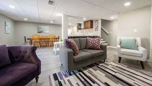Een moderne woonkamer met een grijze bank, een paarse fauteuil en een witte fauteuil. Er is een eethoek met houten stoelen en een tafel, en een keuken met kasten en een aanrecht.