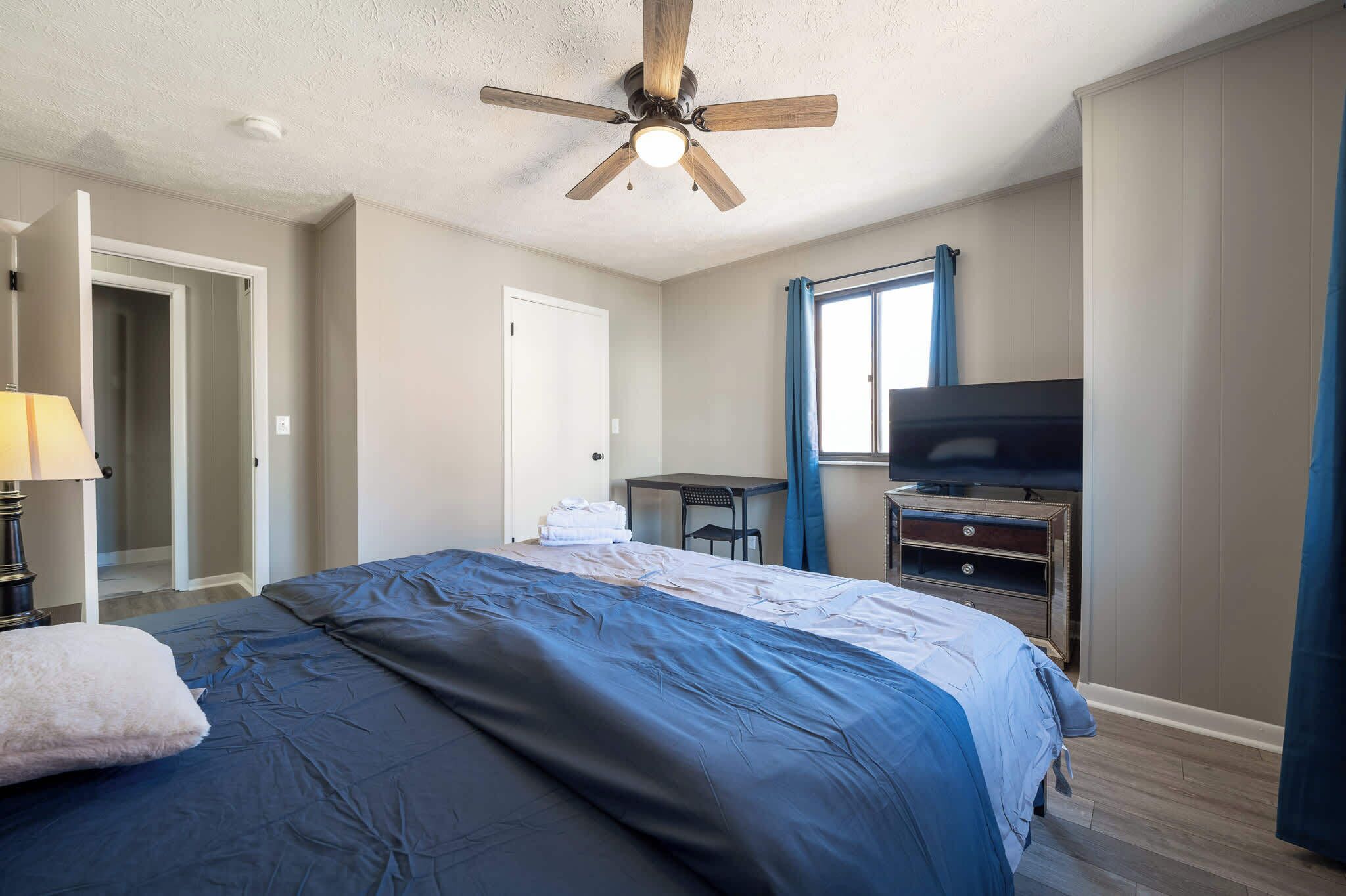 A bedroom with a bed, a ceiling fan, a desk, a television, and a wardrobe.