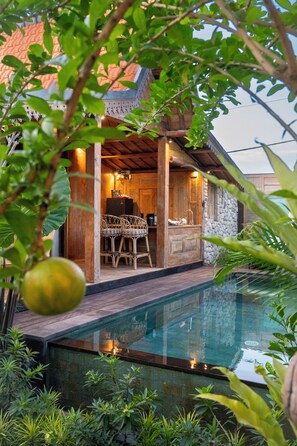 Zona junto a la piscina con terraza de madera, pared de piedra y espacio de descanso con sillas de mimbre.