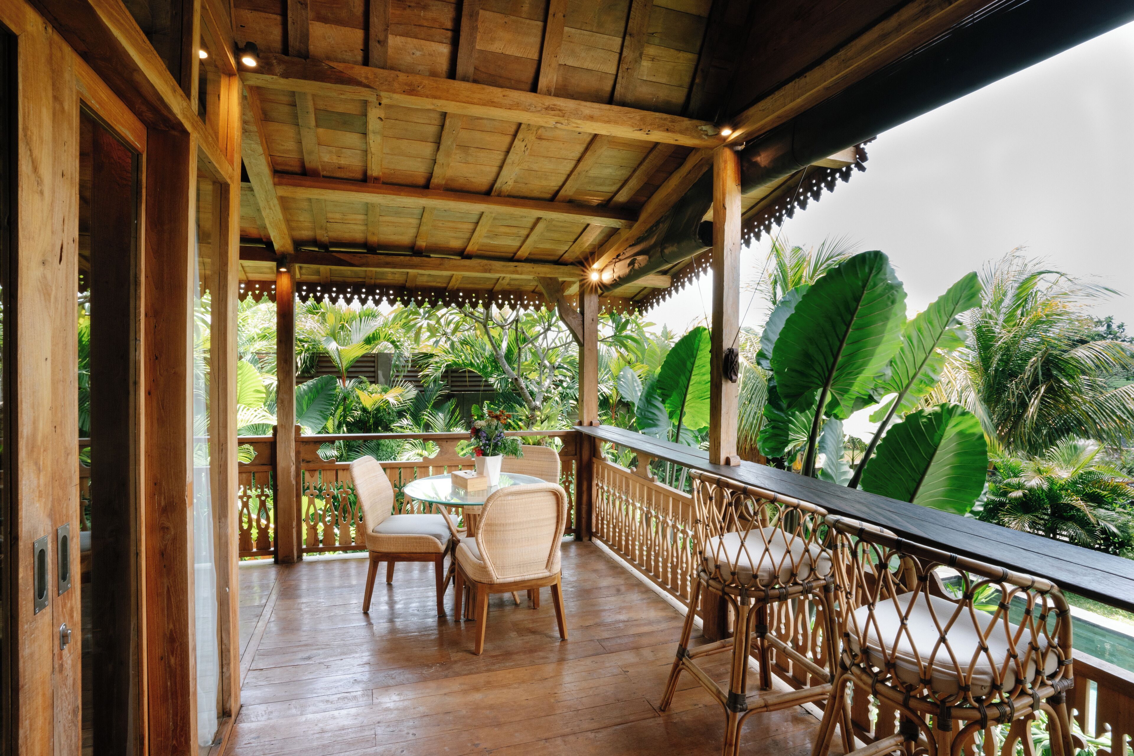 Une terrasse en bois avec une table en verre et des chaises en osier, entourée d’une végétation tropicale luxuriante.