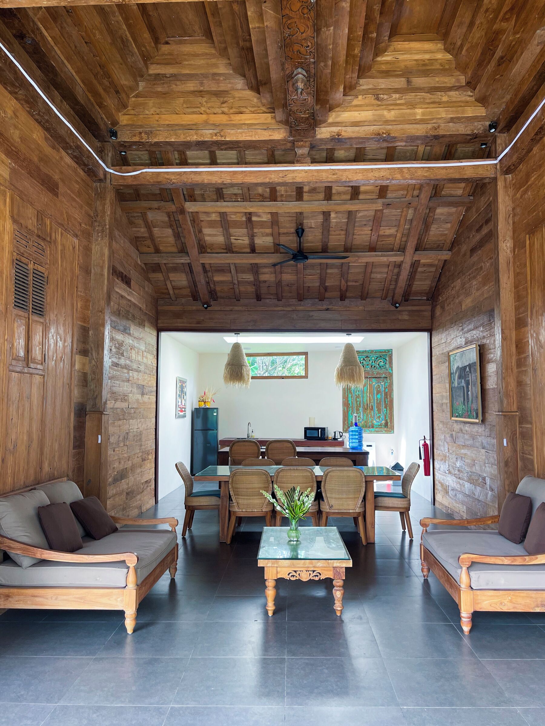 A spacious living room with a high ceiling, wooden beams, a central dining table with chairs, and a sofa set.