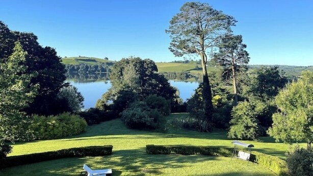 Ein gepflegter Garten mit Seeblick, ordentlich geschnittenen Hecken und einem weißen Stuhl.