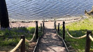 Ein Weg, der zu einem See führt, mit Holzlattenzaun und Seilabsperrung.